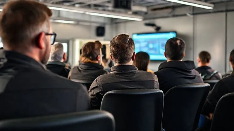 A photorealistic, professional photography shot of a diverse group of commercial truck drivers, both male and female, engaged in a modern safety training session. They are seated in a bright classroom setting, attentively watching a presentation on a screen (not visible in detail). The atmosphere is professional and focused, with cinematic lighting and sharp focus on the participants' expressions. Depth of field, 8K hyper-detailed.